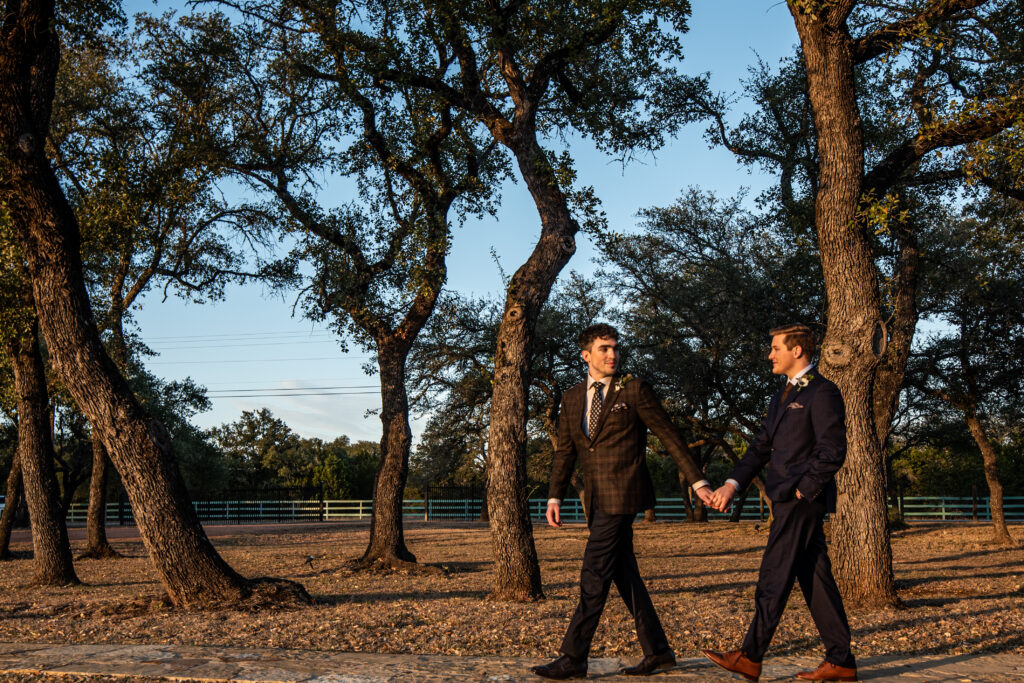 Two grooms holding hands walking into the hill country sunset. One groom looks back at the other.