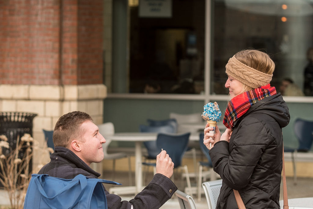 A man kneels for a surprise proposal, offering an engagement ring to a woman holding an ice cream come