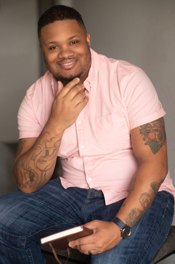 A Black male business owner poses for his headshots, smiling at the camera wearing a pink short-sleeve shirt. 