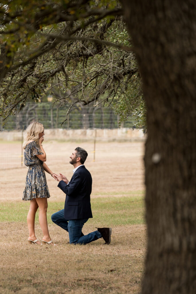 A couple stands at a park, the man down on one knee, proposing to a woman
