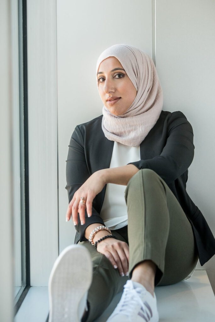 A woman wearing a neutral colored hijab looks into the camera with a soft smile