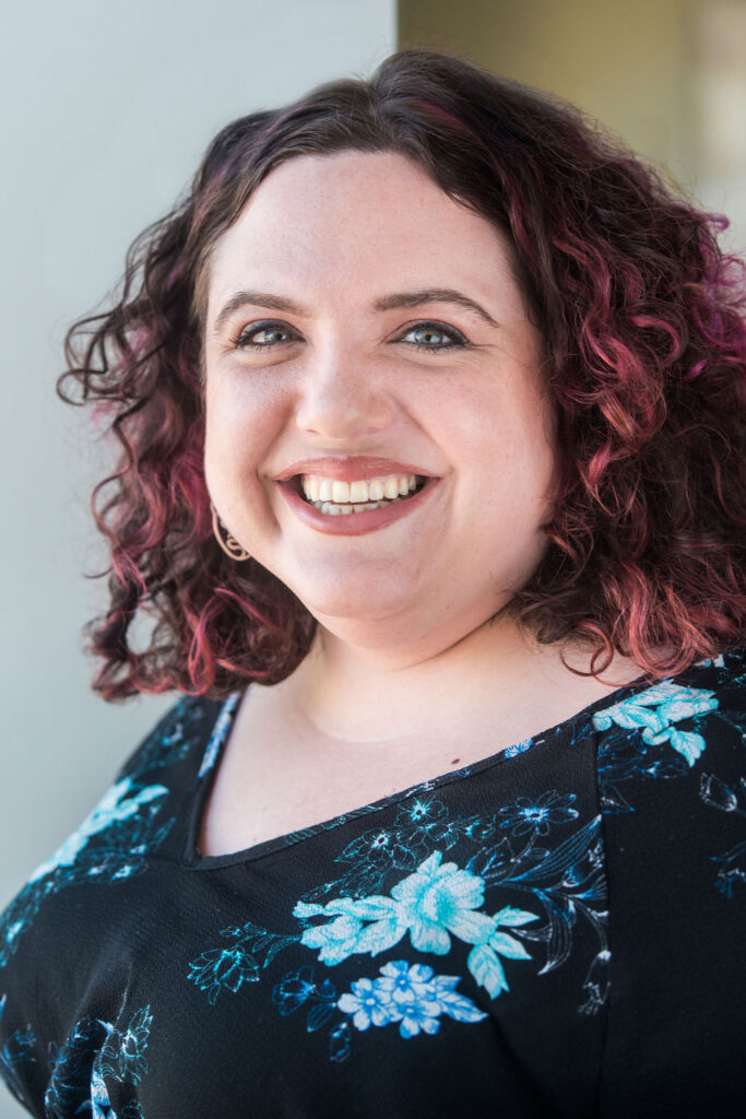 a woman with red-dyed curly hair grins at the camera for her professional headshots session