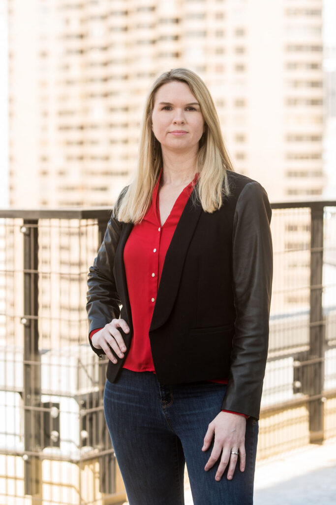 a blonde woman looks to the camera with an urban background wearing a red shirt and black blazer