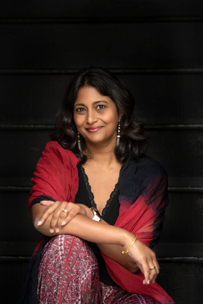 A woman of color poses in front of a black background wearing a red and black outfit