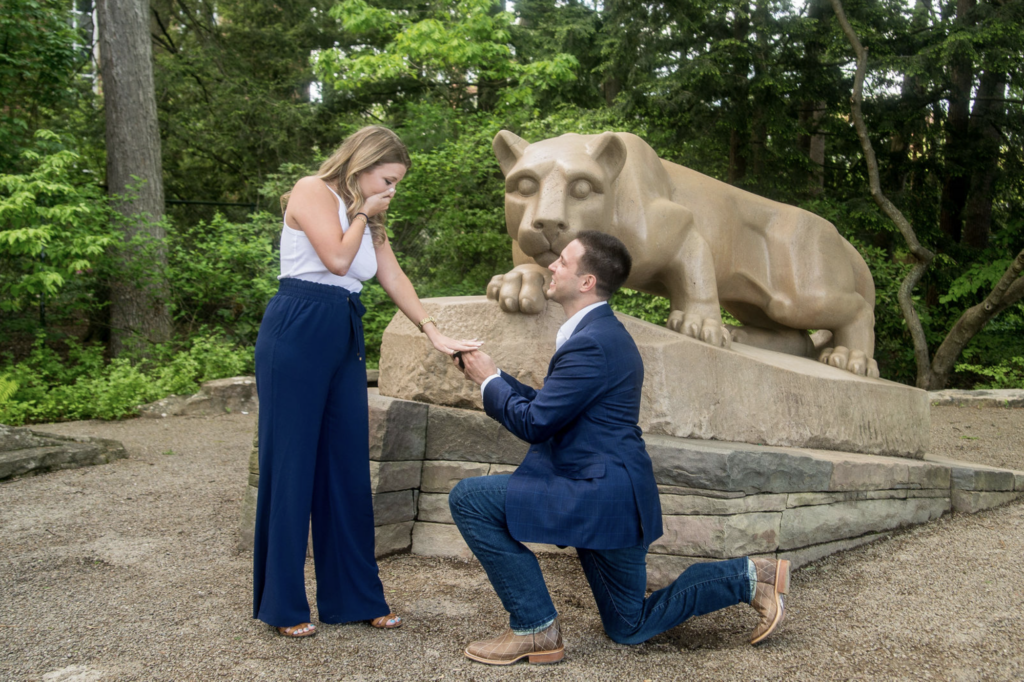 a man and woman stand in a park with a lion statue in the background. He kneels on one knee, offering an engagement ring for his surprise proposal in austin