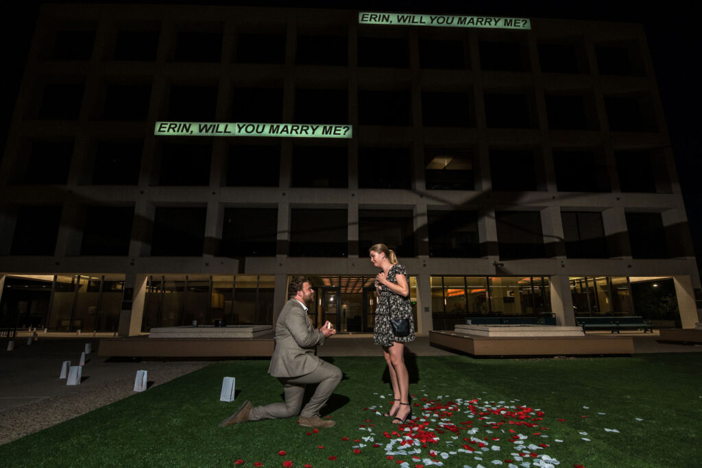 a man plans a surprise proposal in Austin at night, kneeling in front of a woman near a pile of roses