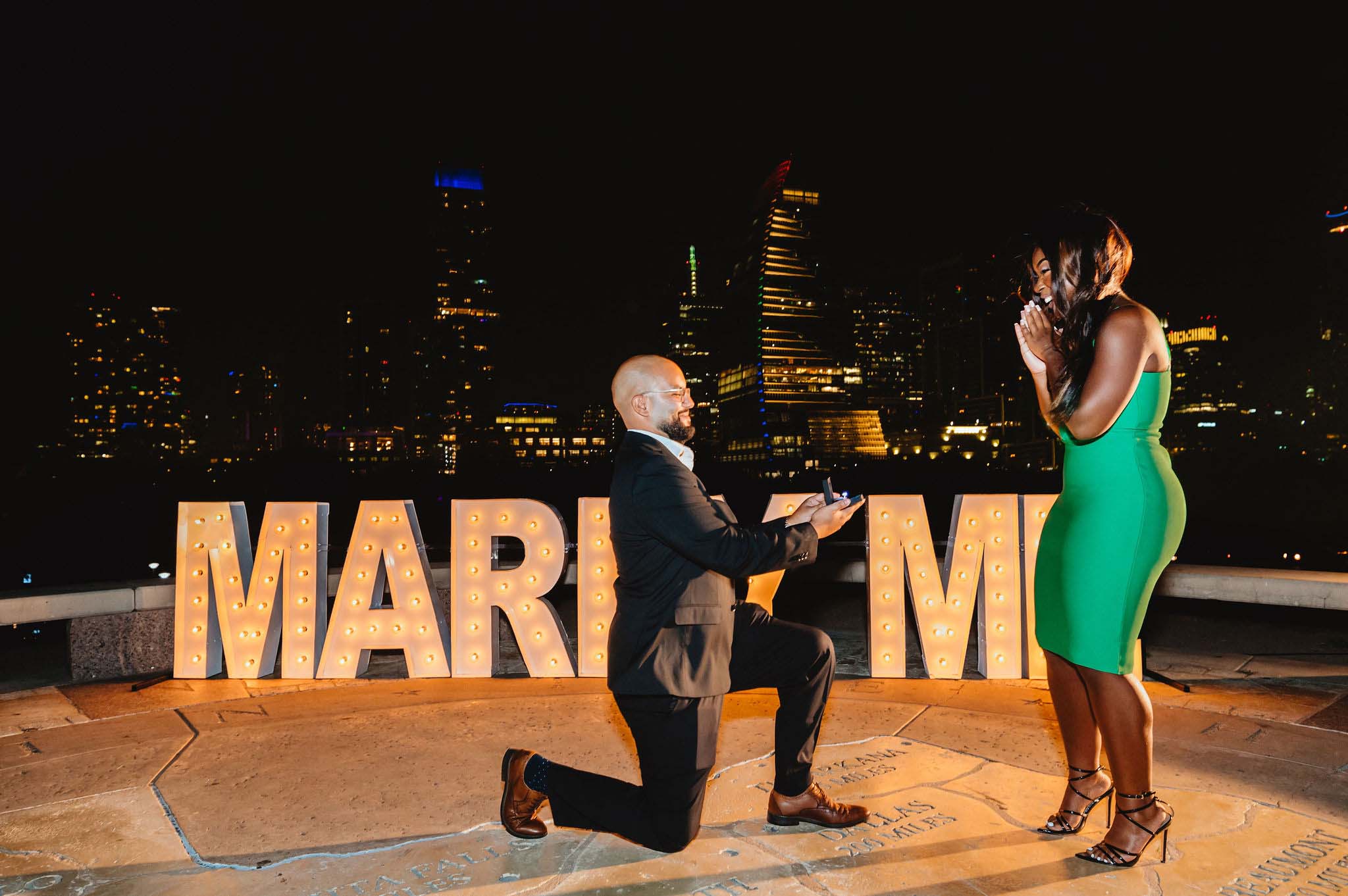 a Black couple is getting engaged in front of a Marry Me sign with downtown Austin in the background
