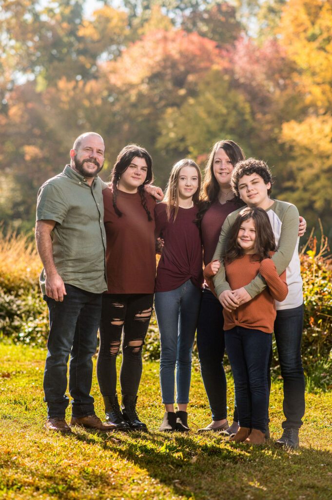 A family gathers for their outdoor family session with fall-colored trees in the background