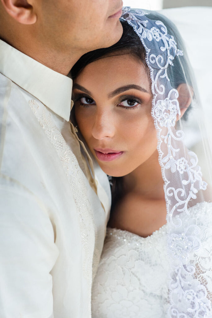 a bride wearing a lace veil leans against her husband while looking at the camera