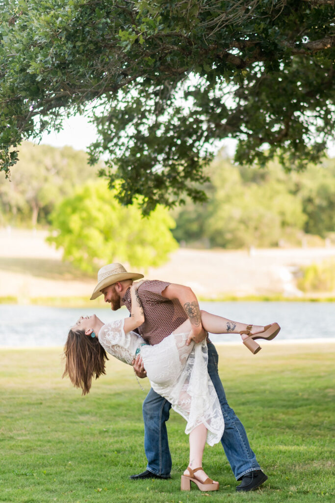 a man dips his fiancee in front of a tree and green lawn
