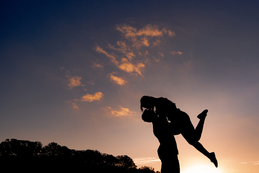 a sunset silhouette of a man lifting his fiance in their air