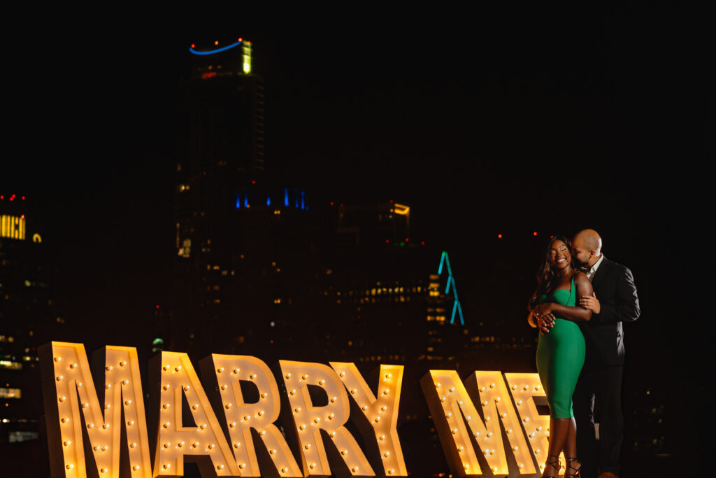 a black couples is holding each other while standing in the Austin skyline. The couple is standing in front of a large marquee that says marry me. 