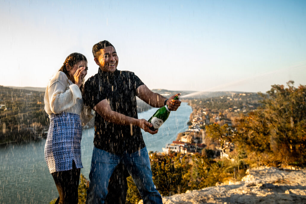 a man and a woman stand on mount bonnell spraying an open bottle of champagne. 