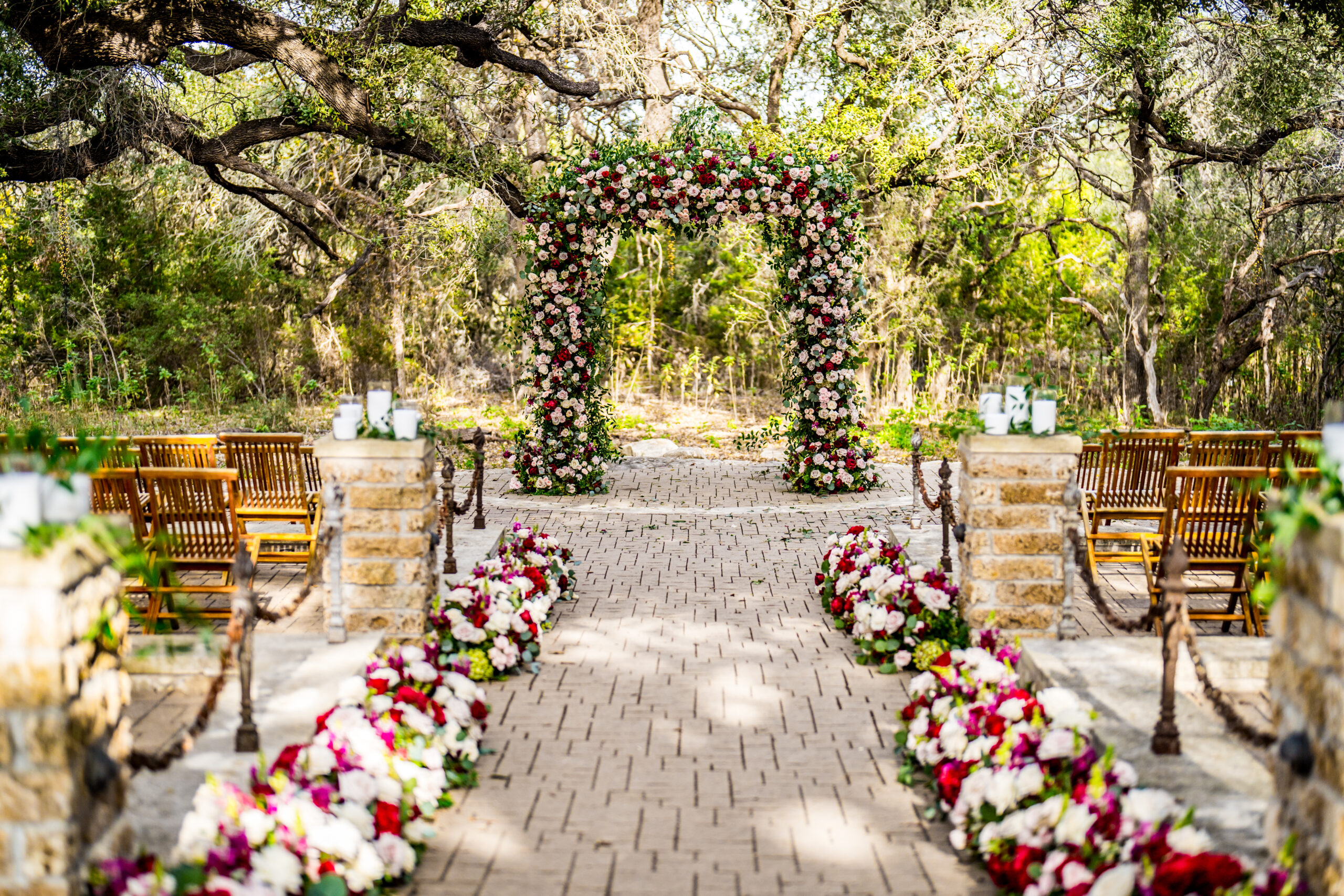 Floral arch at an outdoor wedding ceremony at Sacred Oaks, Camp Lucy, with wooden chairs and oak trees in the background.